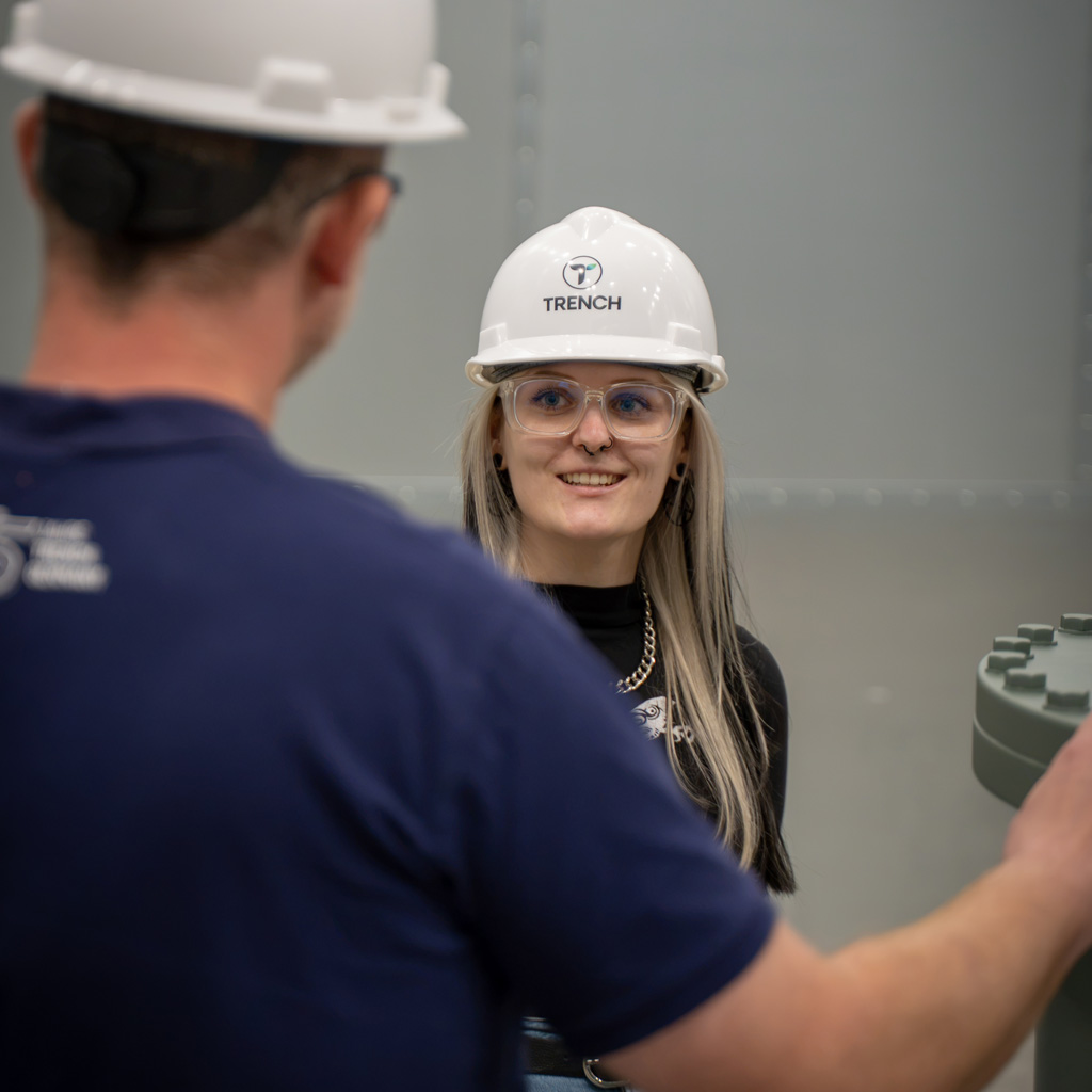 Victoria Bold wearing a Trench-branded safety helmet and talking with a colleague inside the Bamberg facility.
