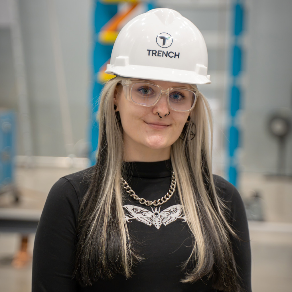 Portrait of Victoria Bold wearing a white Trench-branded safety helmet and protective glasses at the Bamberg facility.